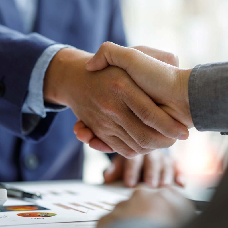 Eine Nahaufnahme eines formellen Handschlags zwischen zwei Personen in Geschäftskleidung. Der Fokus liegt auf den Händen, die sich über einem Schreibtisch mit Dokumenten begegnen, was auf eine erfolgreiche geschäftliche Vereinbarung hinweist.